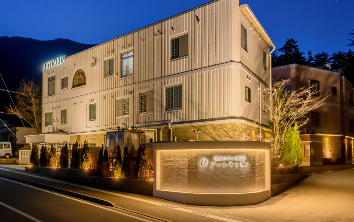温泉ホテル安曇野アートキャビンの南側外観写真。ライトアップされた建物と正面のロゴ壁面が夜空に映える、宿泊予約ページなどに使用される公式イメージ